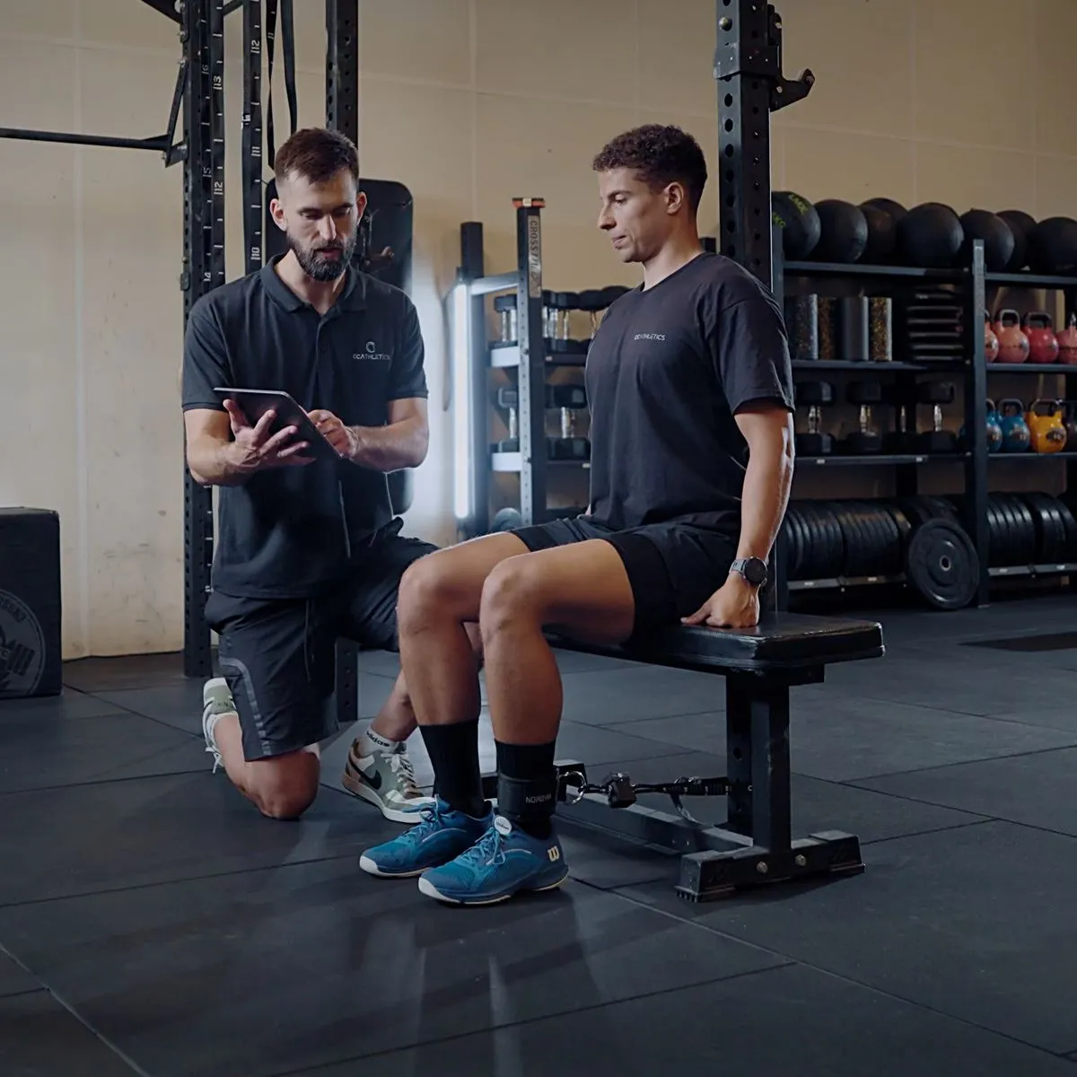 CableMate in use for a seated knee extension test: athlete on a bench with the dynamometer strapped between ankle and bench, while a coach reads the result on a tablet