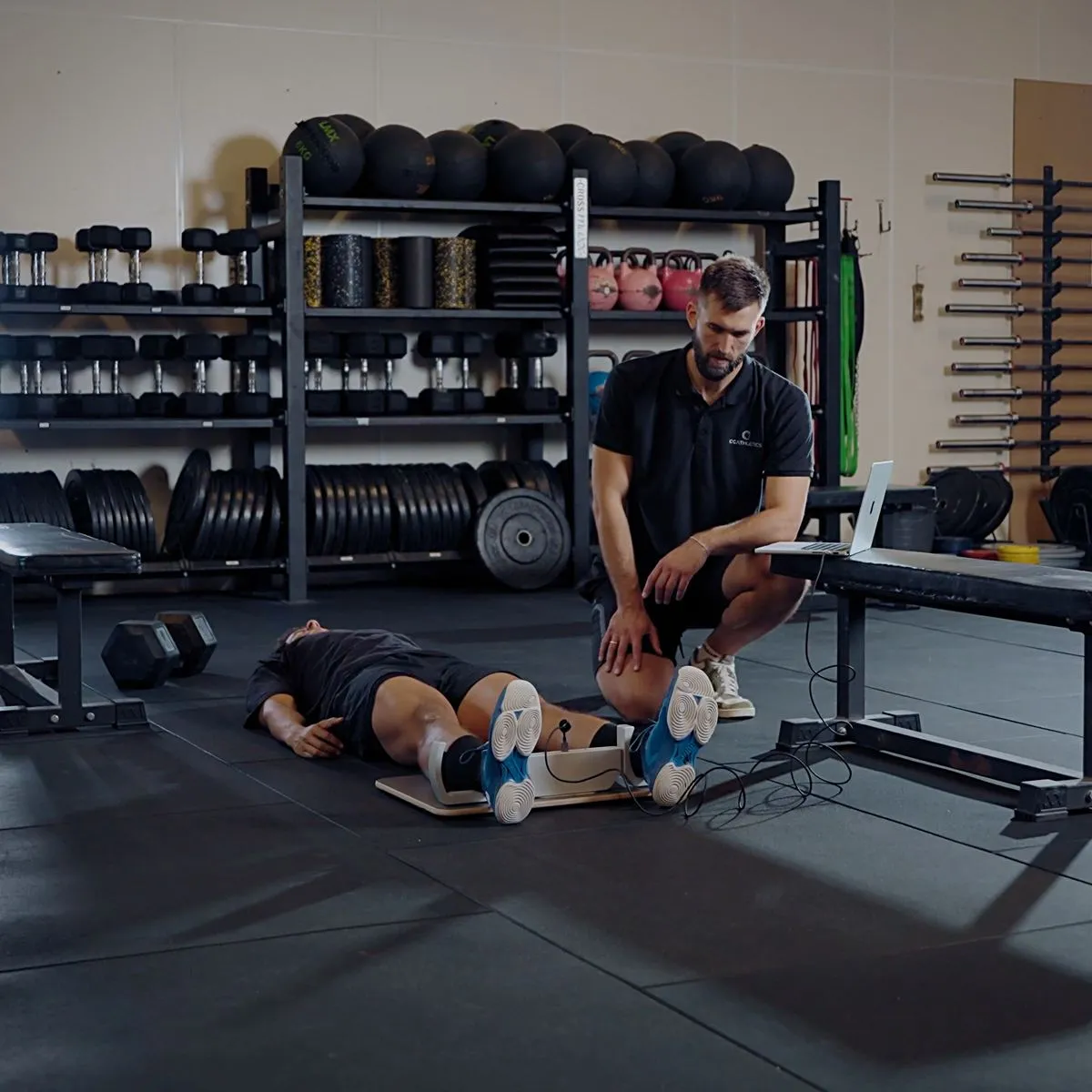 GroinMate in use for an adductor squeeze test: athlete lying on a gym floor with both ankles in the hooks while a coach follows the live reading on a laptop