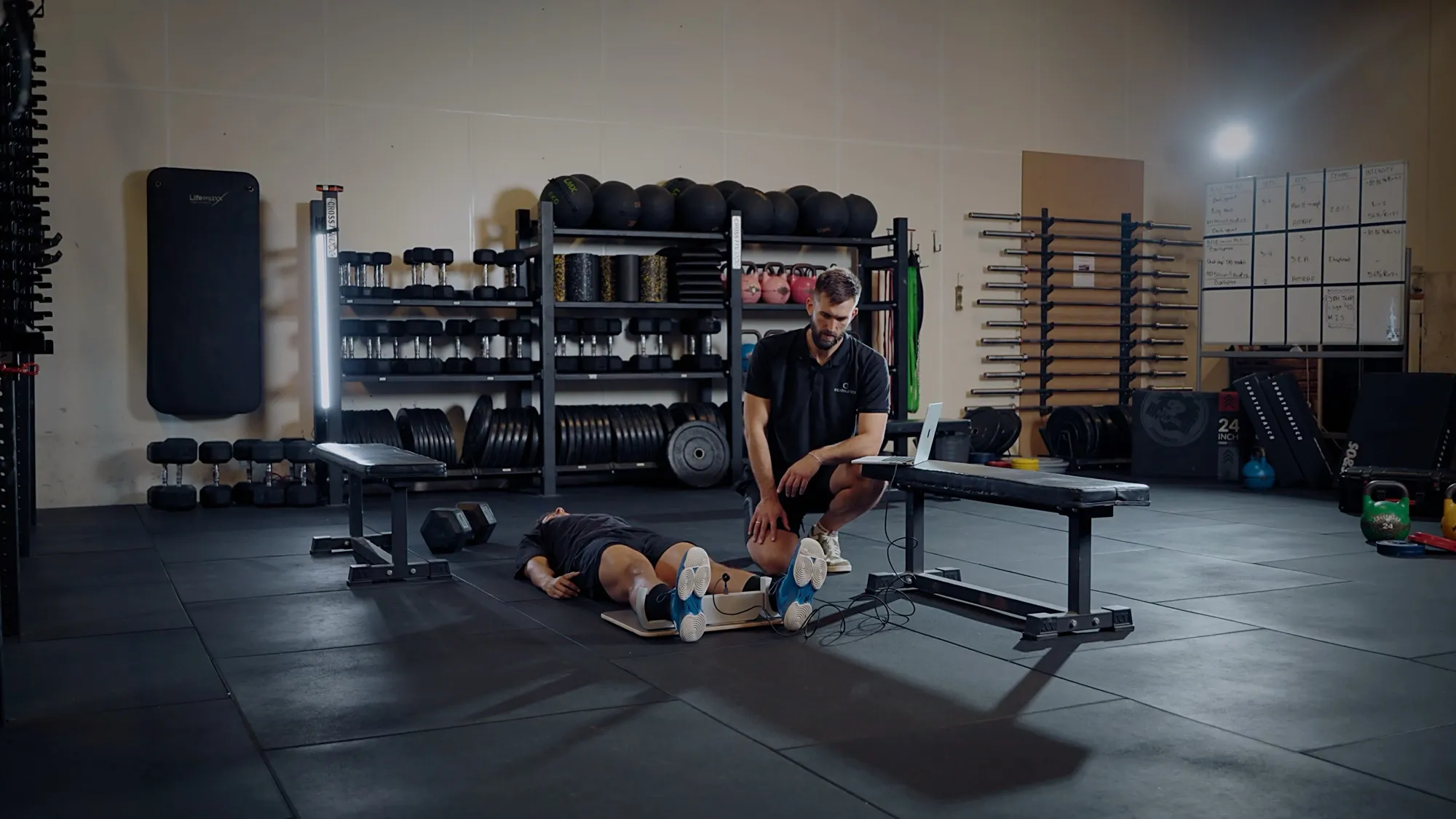 A coach kneels beside an athlete lying on the gym floor performing a GroinMate adductor test, with the results on a laptop